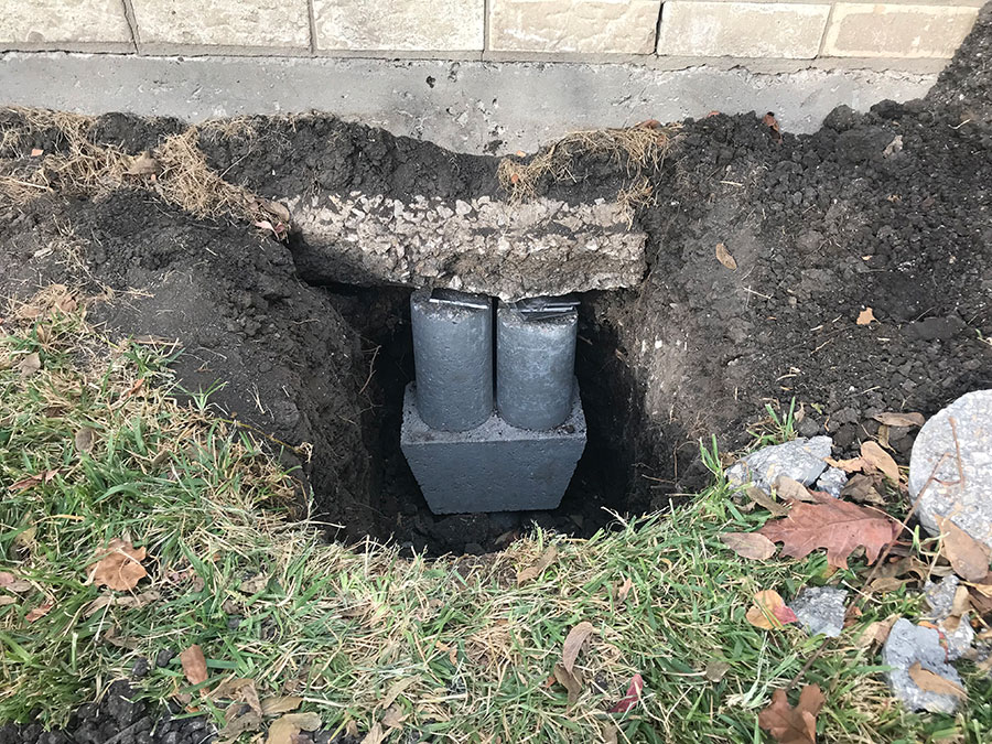 A close-up shot of a foundation repair in progress, showing a deep hole excavated next to a brick building. Inside the hole, two concrete cylinders sit atop a larger base block, supporting the edge of the exposed concrete foundation.