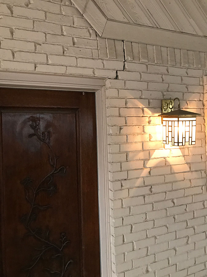 A vertical crack in a white painted brick wall located above the corner of a dark wood door frame. To the right of the crack, a lit mission-style outdoor lantern casts a warm glow on the brickwork.