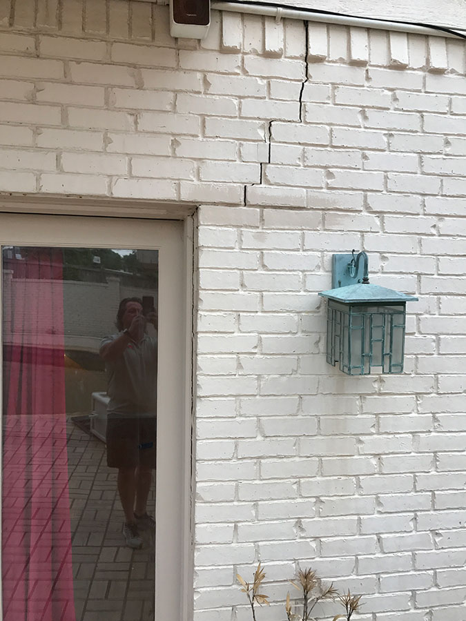 A stair-step crack in a white painted brick wall beginning at the top corner of a white door frame. A decorative green-patina outdoor light fixture is mounted to the right of the crack, which shows visible separation in the mortar and bricks.