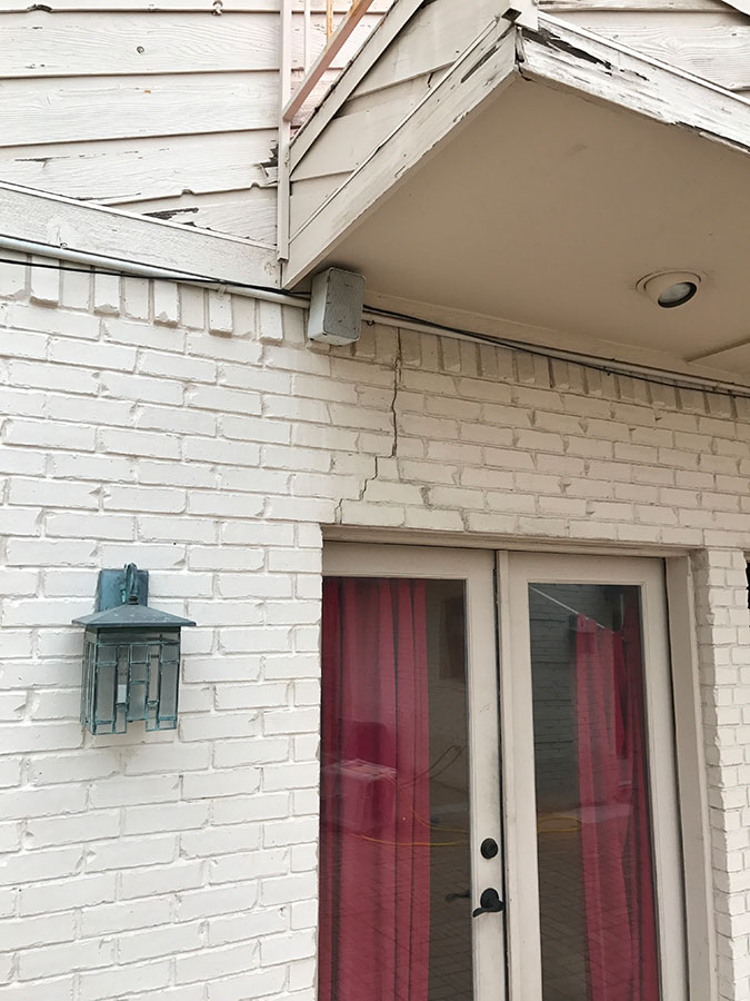 A vertical crack in a white painted brick wall above a set of French doors with pink curtains. The crack extends toward an overhanging balcony with peeling paint. A green-patina decorative lantern is mounted on the wall to the left of the doors.