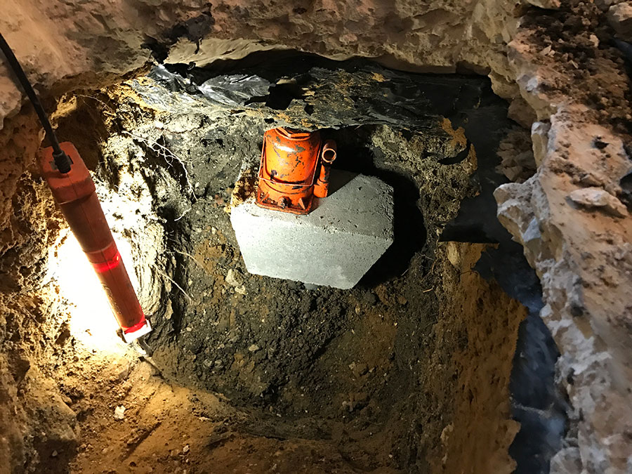 A close-up view inside a foundation repair excavation pit. An orange hydraulic bottle jack sits on a gray concrete pier block, used to level a concrete slab from below. An orange work light on the left side of the frame illuminates the dark soil and the mechanical setup.