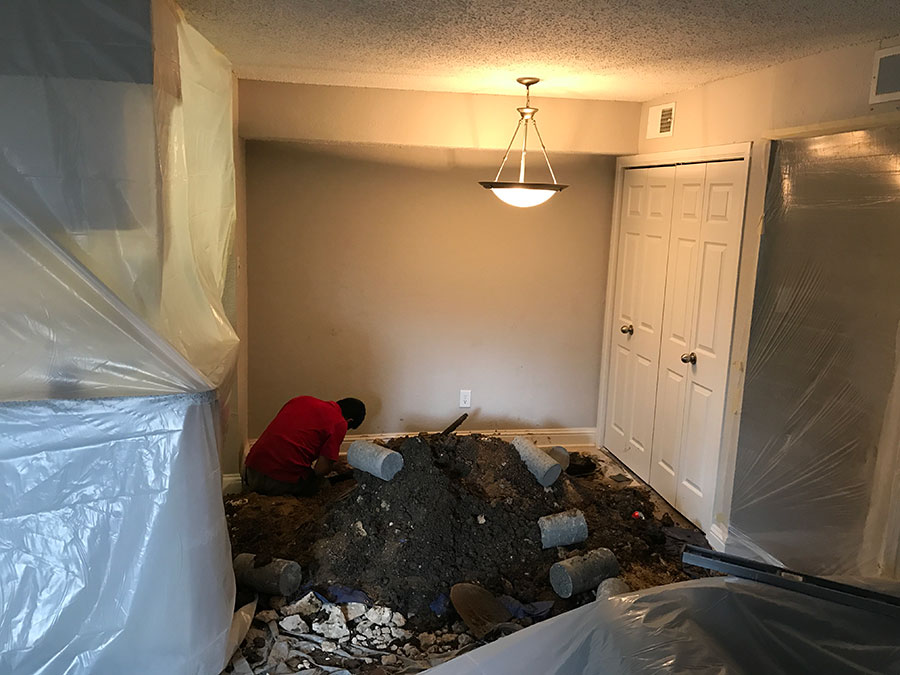 ## Summary of Image An interior room undergoing major foundation repair. A worker in a red shirt is crouched near the wall where a large hole has been excavated through the floor. A significant mound of dark soil and several concrete pier cylinders are piled in the center of the room. To protect the home, the surrounding furniture and walls are draped in clear plastic sheeting. ## Alt Text Interior view of a home during foundation repair. A worker in a red shirt works near an excavation site in the floor, surrounded by a large pile of dirt and concrete cylinders. The room is heavily protected with clear plastic sheeting over the walls and furniture.