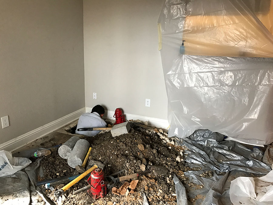 An indoor construction scene where a worker is repairing a foundation from inside a home. A large section of the floor has been removed, creating a hole where the worker is positioned. A mound of dirt, hydraulic jacks, concrete cylinders, and various hand tools sit on plastic sheeting protecting the remaining carpet. To the right, furniture is covered in clear plastic for protection.