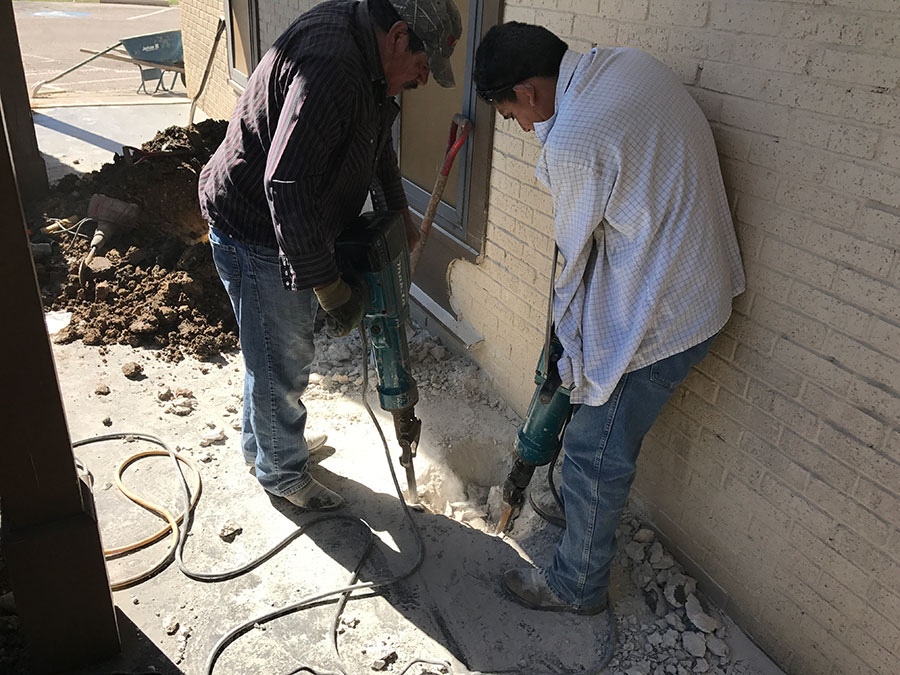Two workers using electric jackhammers to break through a concrete walkway alongside a light-colored brick building. Debris and a pile of excavated dirt are visible in the background, indicating ongoing foundation or utility repair work.