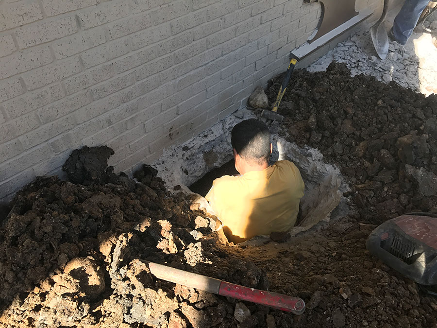 A worker in a yellow shirt is partially inside a deep excavation hole at the base of a light-colored brick wall. The site is surrounded by large mounds of dark soil and construction tools, including a shovel and a sledgehammer, indicating an active foundation repair or utility project.