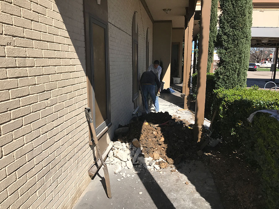 Construction workers excavating a hole at the base of a light-colored brick building's foundation. A pile of dark soil and chunks of broken concrete sit on the walkway next to the dig site, with a shovel leaning against the wall in the foreground.