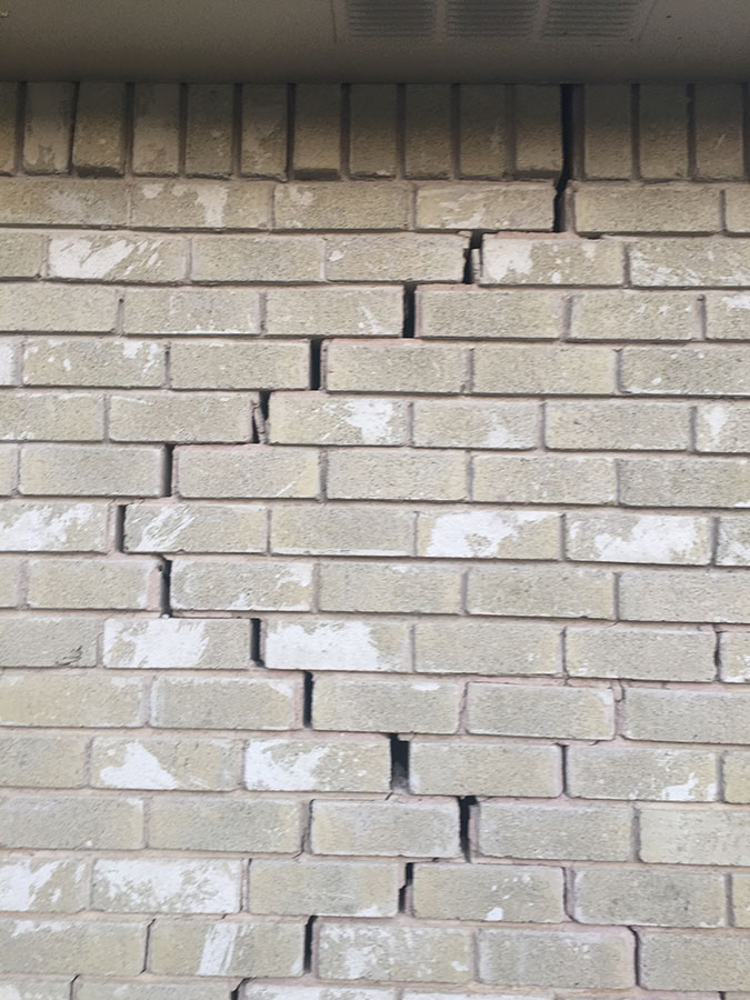 A light-colored brick wall featuring a significant stair-step crack that follows the mortar lines diagonally from the top right toward the bottom left. The separation is wide enough to see dark gaps between the bricks, indicating potential foundation movement or structural settling.