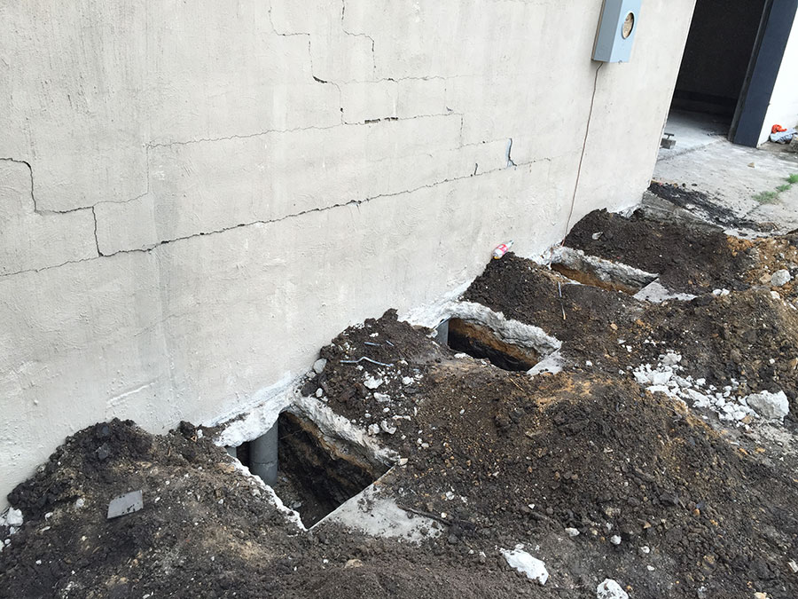 A stucco wall showing multiple horizontal and stair-step cracks, with several deep rectangular holes excavated along the base of the foundation for repair work. Steel piers or supports are visible inside the holes, surrounded by mounds of displaced dirt.