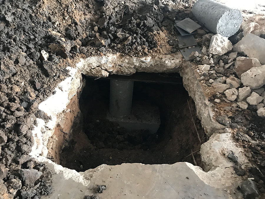 A top-down view of a foundation piering installation, showing a deep rectangular hole cut through a concrete slab. Inside the excavation, a concrete support cylinder is positioned to stabilize the structure, surrounded by loose soil and rubble from the dig.
