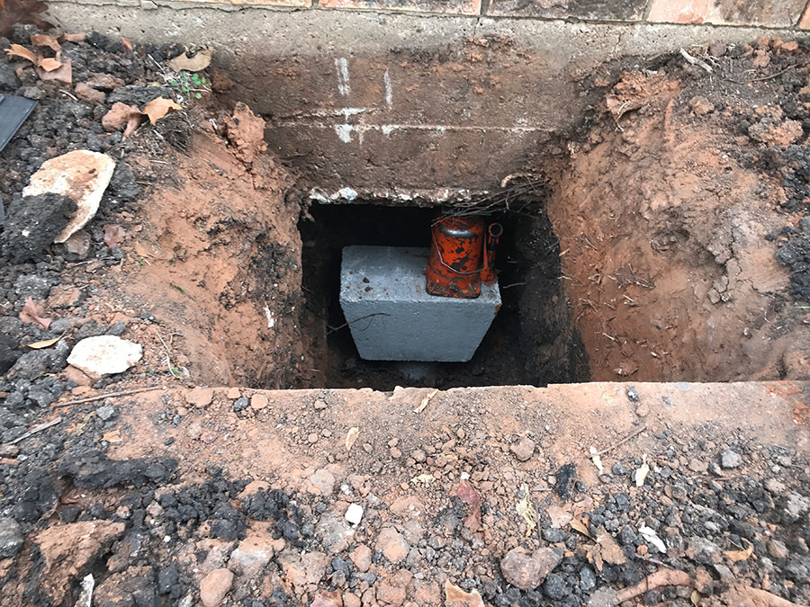 A top-down view of a foundation repair excavation next to a brick wall. Inside the dirt pit, an orange hydraulic jack is positioned on a concrete pier block to level and support the building's foundation.