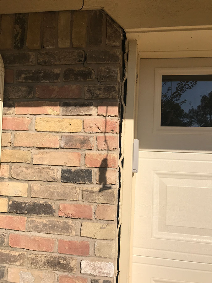 Exterior shot of a brick wall pulling away from a white garage door frame, creating a wide, dark gap. A stair-step crack is also visible in the mortar of the bricks near the top of the frame, and a small crack appears in the overhead soffit.