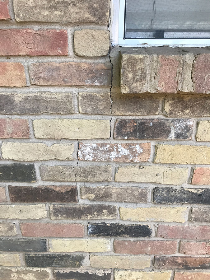 A vertical crack in a brick wall with varied earth-tone bricks, starting from the corner of a window sill and extending downward. The crack clearly splits several bricks and mortar joints, indicating structural shifting.