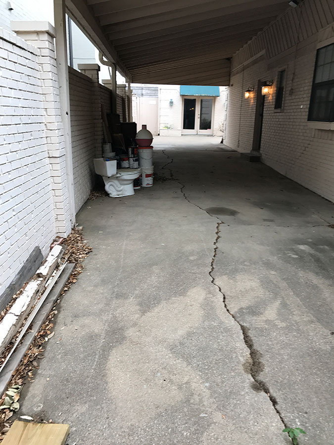 An outdoor covered driveway with a long, prominent crack running through the concrete, flanked by a white brick wall and a building with various items stored along the side, including a toilet and paint buckets.