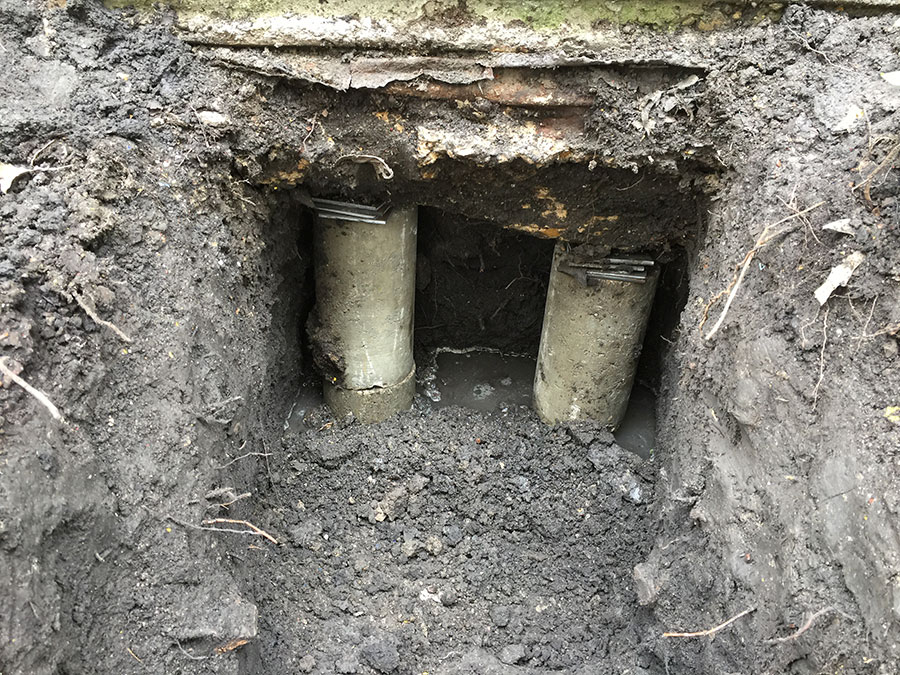 ## Summary of Image This image provides a top-down view into an excavation pit for a **foundation repair**. Two concrete pressed pilings (piers) have been installed deep into the soil to support the structure's foundation. Steel shims are visible on top of the concrete cylinders, used to bridge the gap and provide precise leveling between the new piers and the existing concrete beam. Some standing water is visible at the bottom of the muddy pit. ## Alt Text A deep excavation hole showing two concrete foundation piers installed under a concrete beam. Small steel shims are stacked on top of the piers to level the foundation. The surrounding soil is dark and moist, with a small amount of water at the base of the pit.